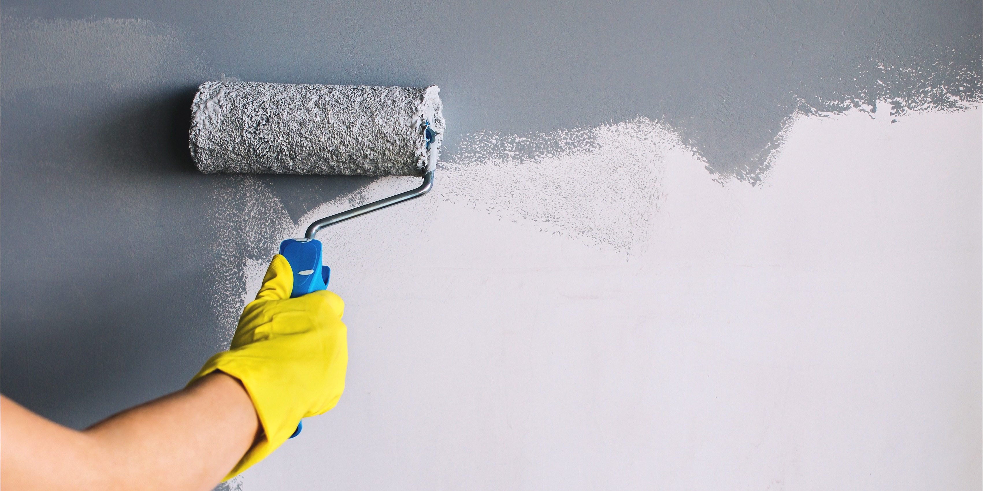 Hand in yellow glove painting wall in gray color with a roller brush.