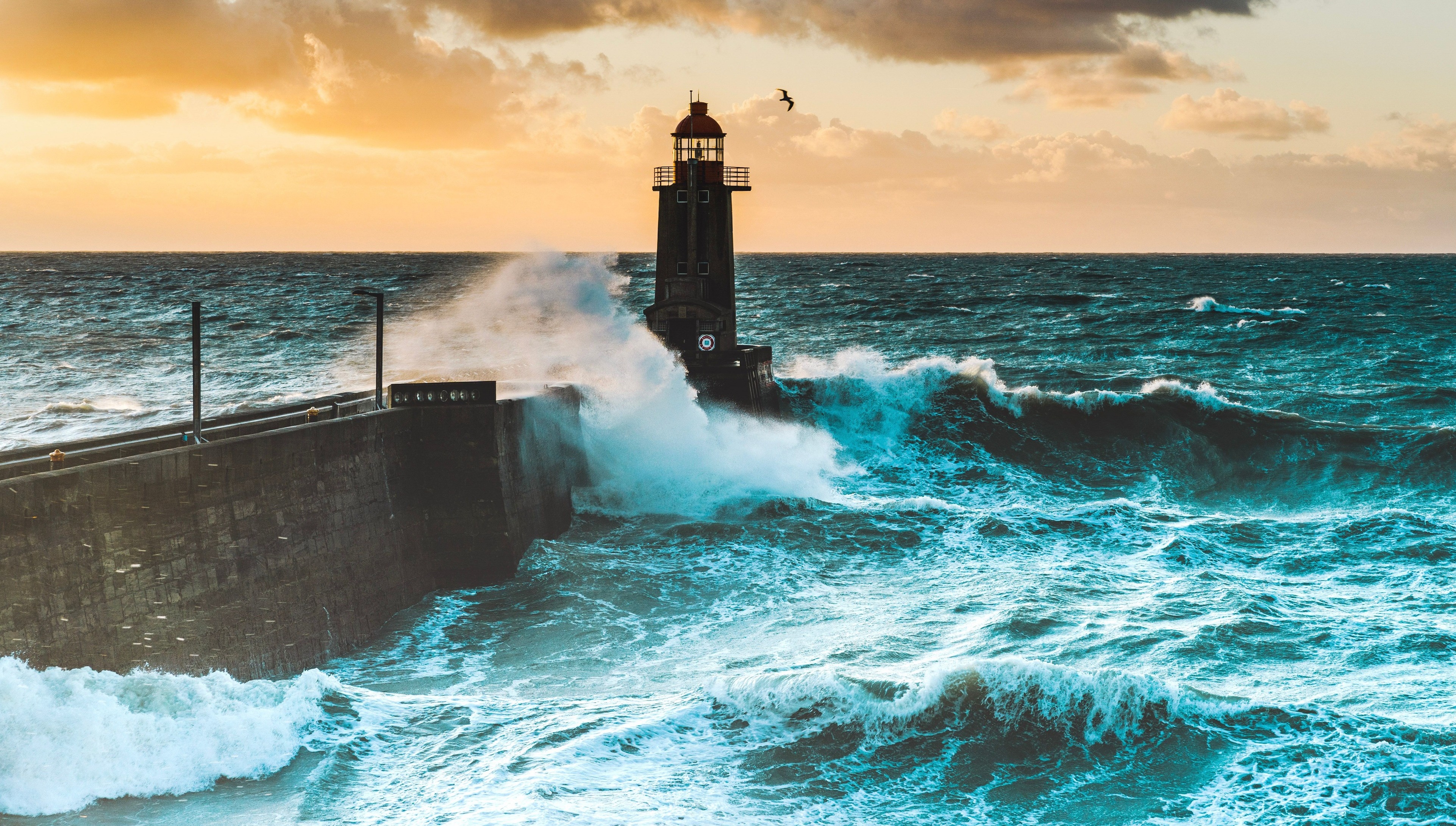 A large waves crash against the stone tower of the lighthouse at high tide at sunset